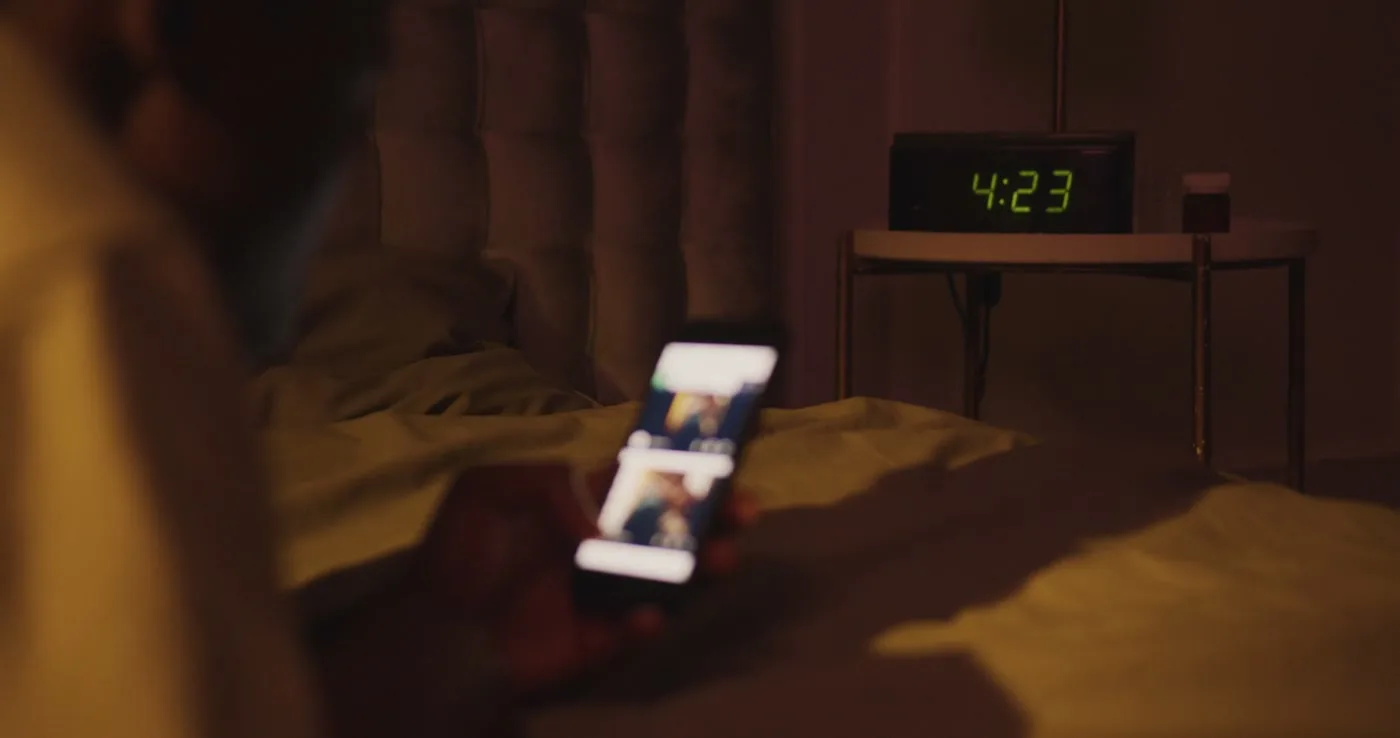 A phone glows on a bed beside an alarm clock reading 4:23.