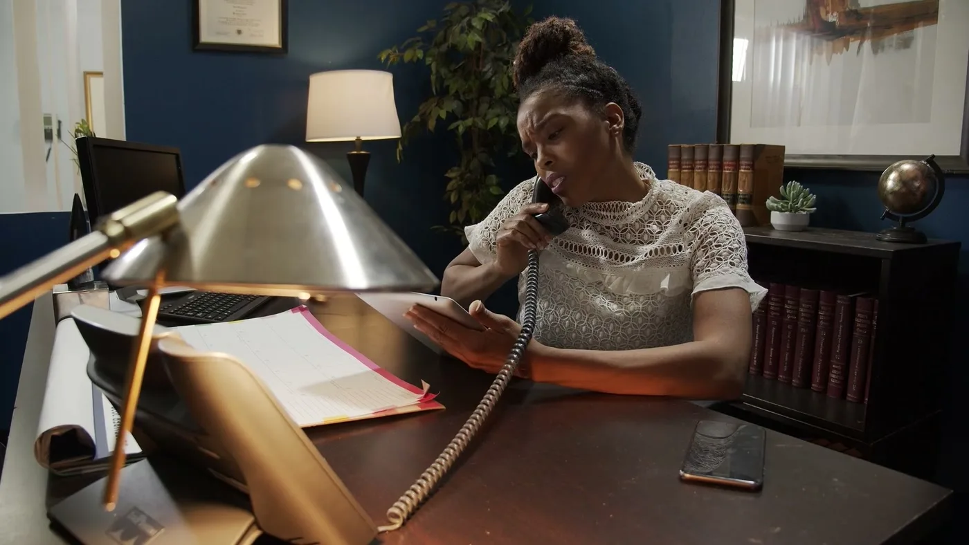 A woman at a desk takes a call on a corded phone, files open in front of her.