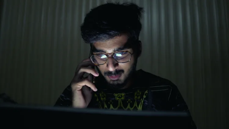 A man answers a phone call in the dark, his face lit by a laptop screen.