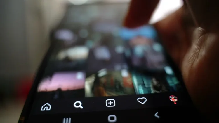 A thumb scrolls a social media grid on a phone in the dark.